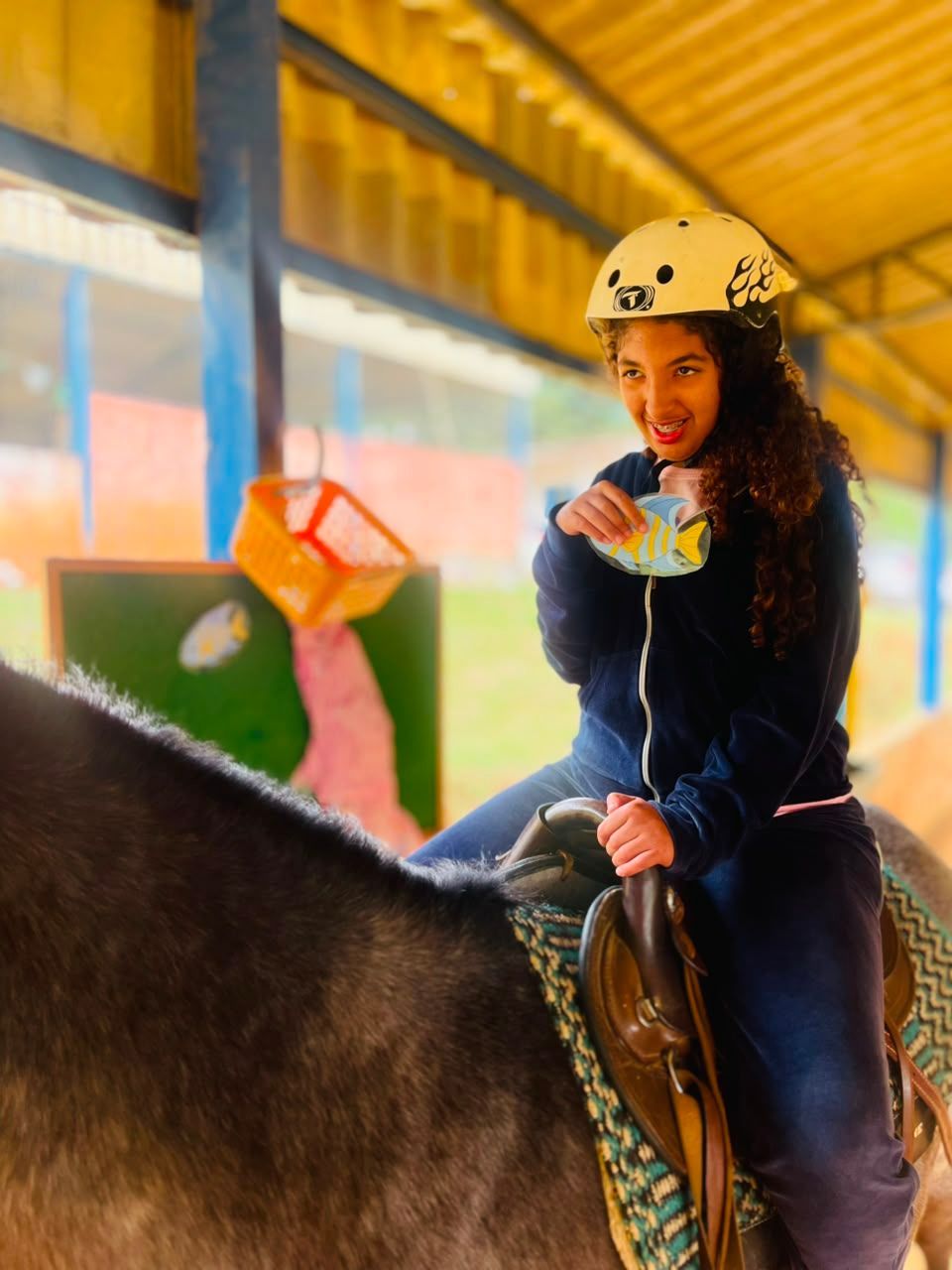 Jovem de capacete sorrindo, montada no cavalo, no picadeiro coberto.