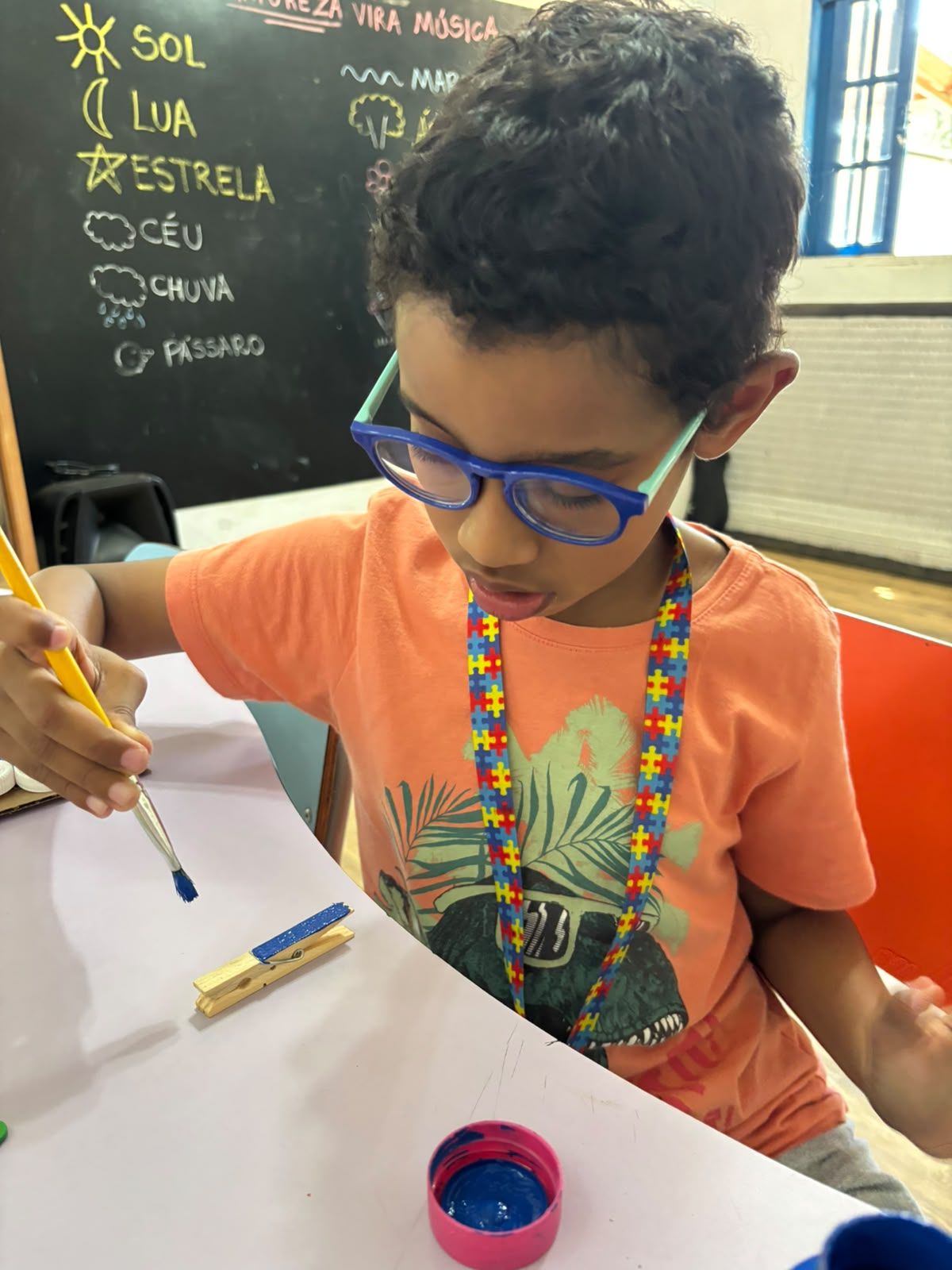 Menino de óculos azuis pintando com pincel numa mesa branca.