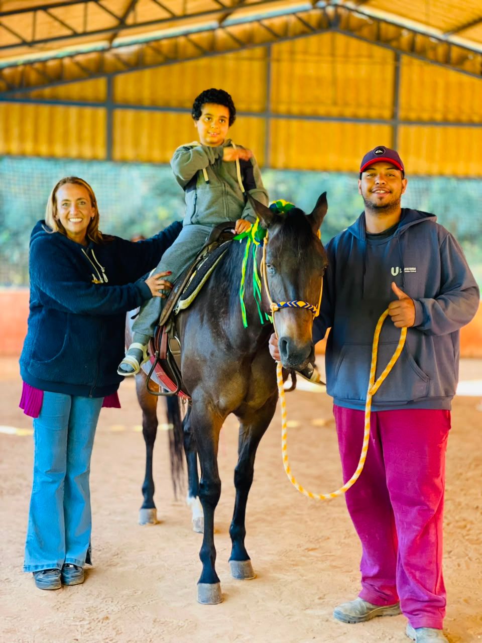 Dois profissionais ao lado do cavalo, com um menino montado e sorrindo.