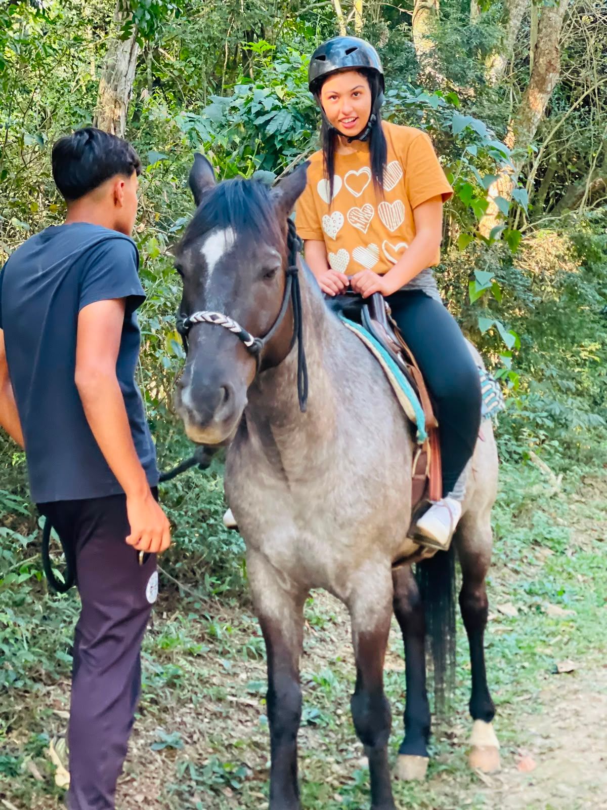 Jovem sorrindo, montada num cavalo tordilho, na trilha da mata.